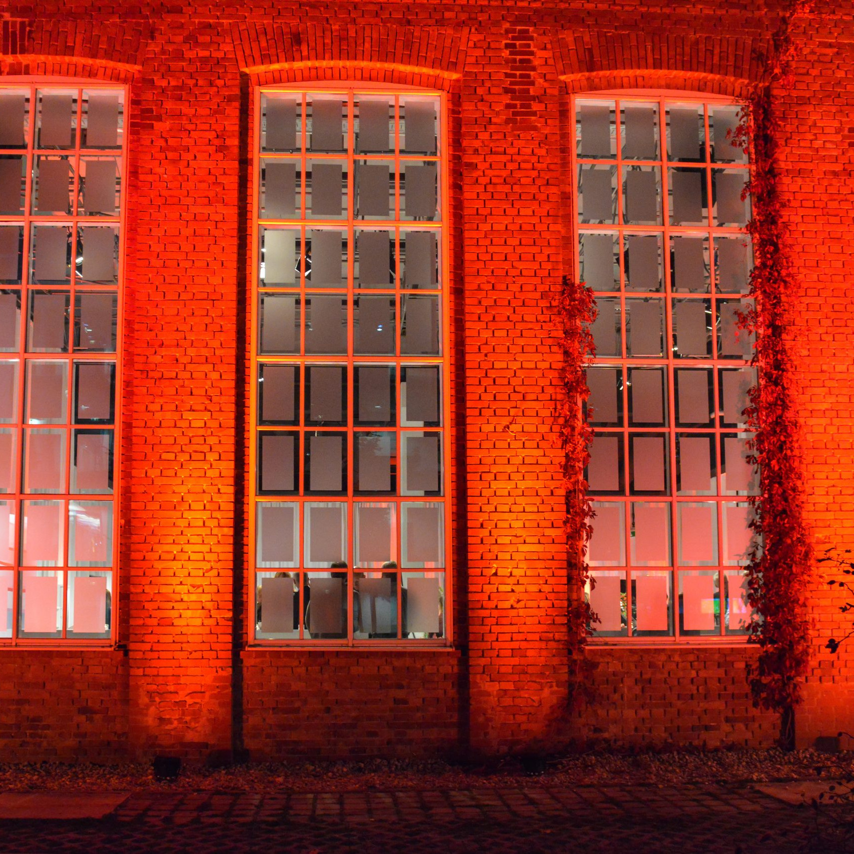 Aliud Pharma: Altes Backsteingebäude mit großen Fensterfronten in rot beleuchtet.