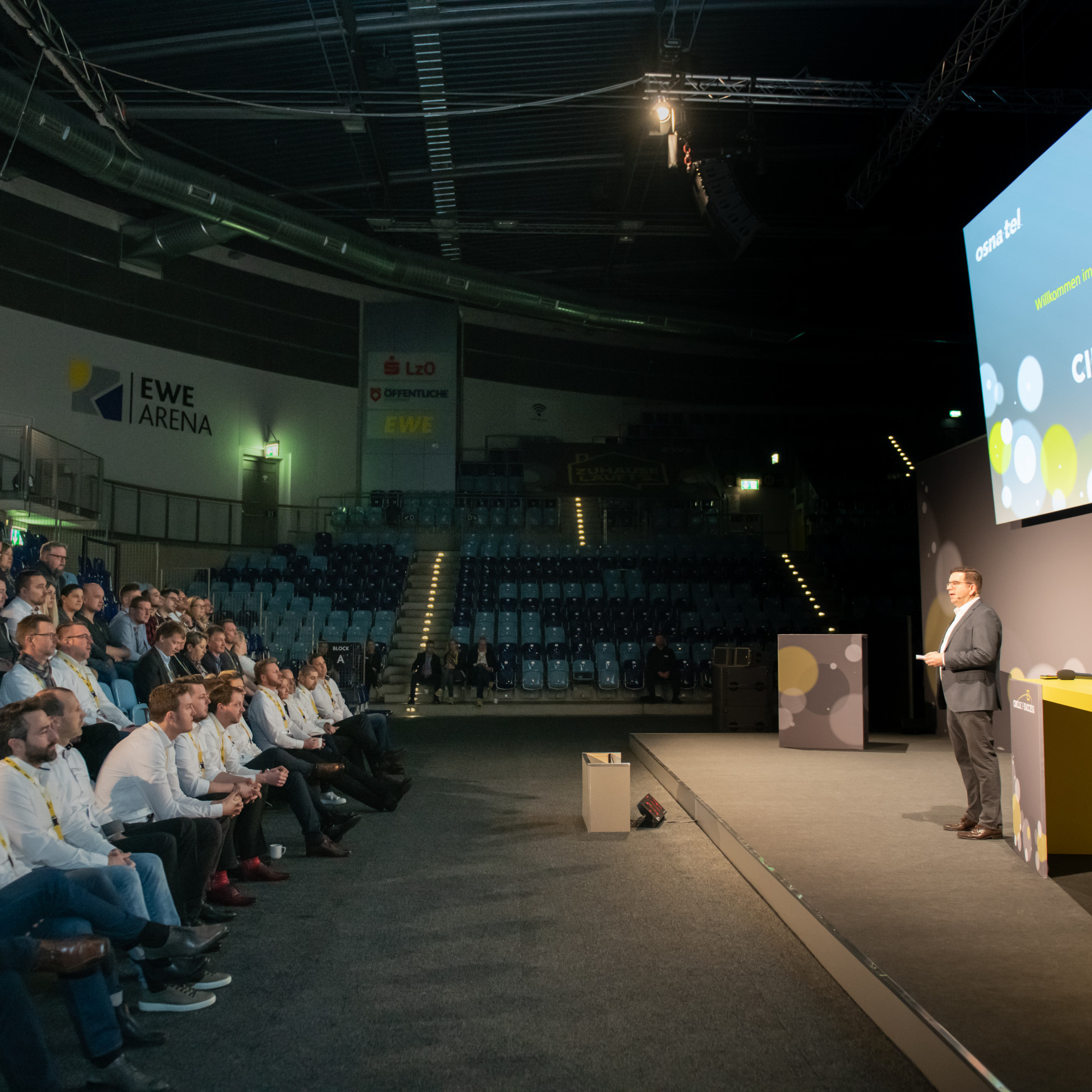 EWE Tel Partnerforum: Seitlicher Einblick in den Tagungsbereich und auf die Bühne. Hier steht ein Redner und spricht. Links das Publikum.