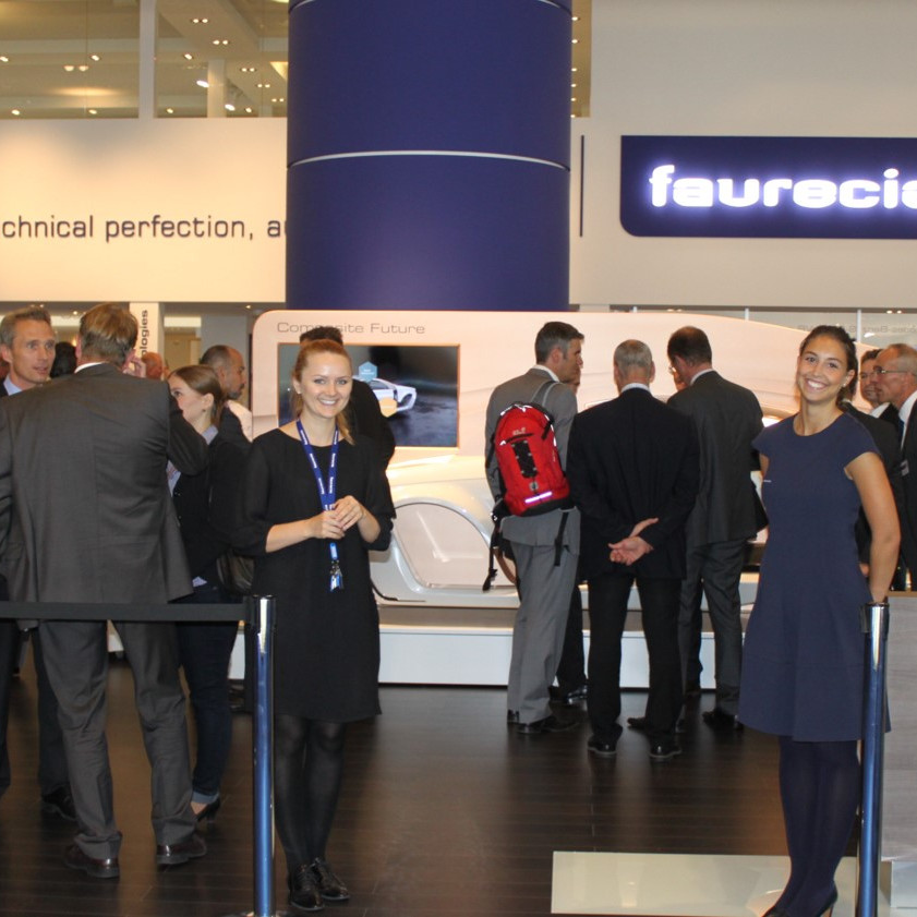 Faurecia Messe: Zwei Messehostessen begrüssen die Gäste des Messestands. Auf dem Messestand sind jede menge Menschen