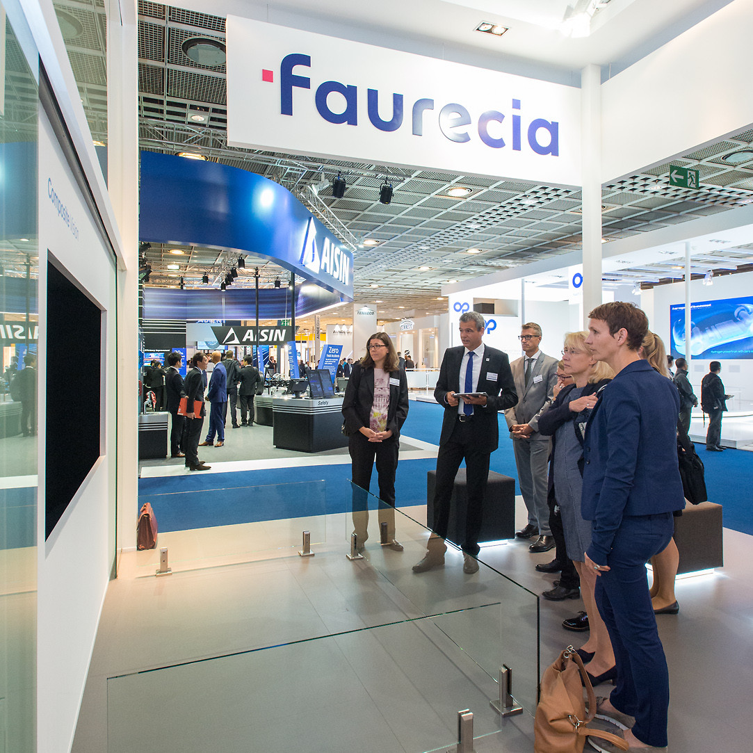 Faurecia Messe: Eine kleine Gruppe Menschen steht vor einem Monitor und schaut sich einen Film an.