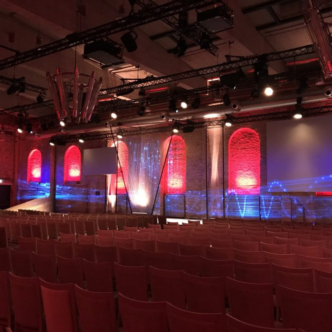 Fraunhofer Jahrestagung: Blich von schräg rechts hinten durch den Saal. Die Wände sind rot illuminiert und vor der Bühne stehen weiße Stühle.