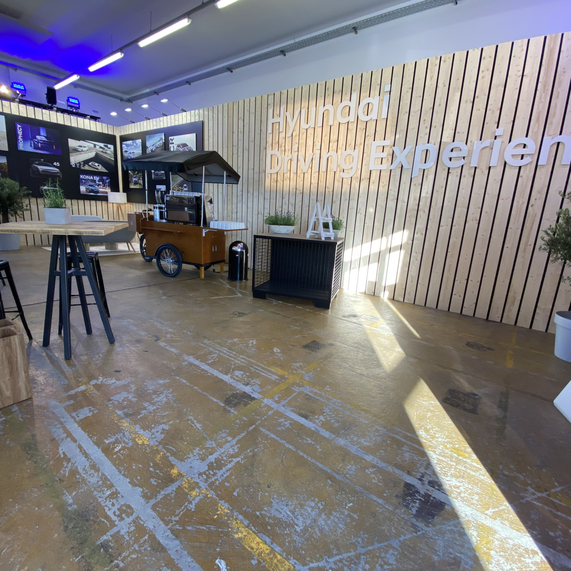 Hyundai Driving Experience: Die Wände des Raumes sind mit Holz verkleidet. Vor der Wand steht ein kleiner Marktstand bei dem es Kaffee und Tee gibt.