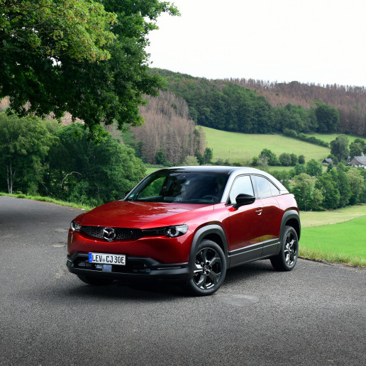 Mazda MX30 Presse Launch: Ein roter Mazda MX30 steht mitten in der Landschaft auf einem Schotterweg, im Hintergrund ist Wald zu sehen.