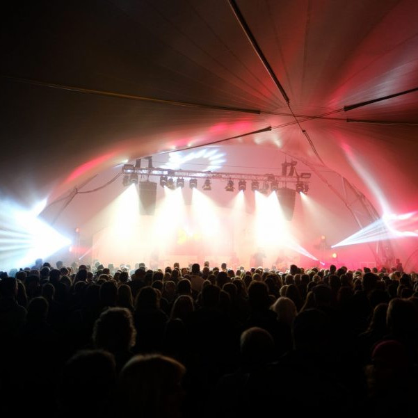 OLB 150 Jahre Festival: Blick in das volle Zelt auf die Bühne. Jede Menge Menschen feiern vor der Bühne, die hell erleuchtet ist.