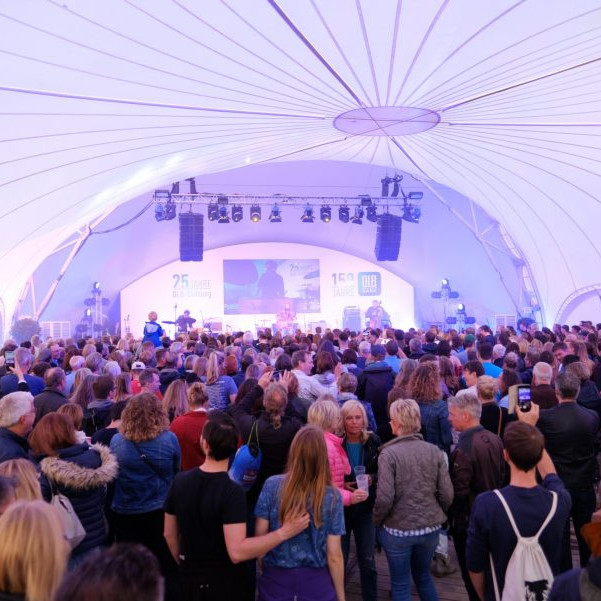 OLB 150 Jahre Festival:Blick von hinten durch das Zelt auf die Bühne.Vor der Bühne stehen jede Mendge Menschen
